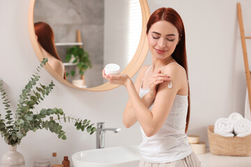 Beautiful young woman applying body cream onto shoulder in bathroom, space for text