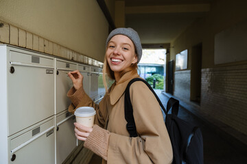 Smiling woman opening mailbox with key and drinking coffee while standing outdoors