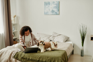 Wall Mural - Cozy portrait of black young woman relaxing at home sitting on bed with cute pet dog and using laptop, copy space