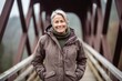 Medium shot portrait photography of a grinning mature woman wearing a durable parka against a rustic bridge background. With generative AI technology
