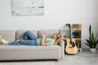 side view of happy young woman with blonde and short hair, bangs and eyeglasses using smartphone while resting on comfortable couch near guitar in modern living room with painting on wall and plants