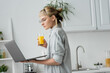 © LIGHTFIELD STUDIOS - young woman in eyeglasses, short hair and bangs holding glass of fresh orange juice and using laptop while working from home in white and modern kitchen, blurred background, freelancer