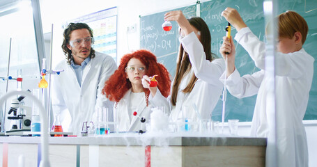 Male and female pupils wearing safety glasses and lab coats mixing liquids in test tubes under close supervision of schoolmaster. Learning process with experiments at modern school.
