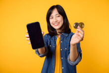 Happy Young Asian Woman Wearing Yellow T-shirt Denim Shirt Holding Digital Crypto Currency Coin And Smartphone Isolated On Yellow Background. Digital Currency Financial Concept.