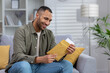 © Tetiana - A smiling young African-American man is sitting on the sofa at home and opens an envelope in which he received a letter, invitation, message