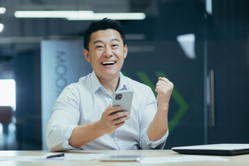 Portrait of a young Asian businessman sitting in the office and holding the phone, received a message of good news, financial successful deal. Shows a victory gesture and rejoices at the camera.
