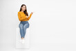 © comzeal - Young Asian woman in yellow shirt sitting and pointing to empty copy space isolated on white background, Full body composition