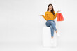 © comzeal - Asian woman sitting on white box and holding red shopping bags isolated on white background, Shopper or shopaholic concept