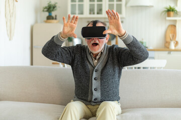 Excited mature senior man wearing using virtual reality metaverse VR glasses headset at home. Grandfather touching air during VR experience on virtual reality helmet. Simulation hi-tech videogame