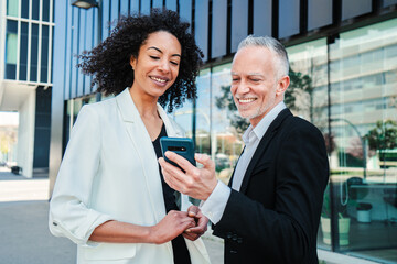 Two business people using a cellphone to watch the corporate shares. Businessman showing his smartphone app to his businesswoman partner. Corporate colleagues wearing formal suit talking at workplace