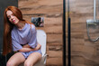 Portrait of sick young woman suffering from ache and holding painful stomach sitting on toilet bowl at home bathroom. Redhead female during menstruation feeling stomach ache sitting on toilet diarrhea