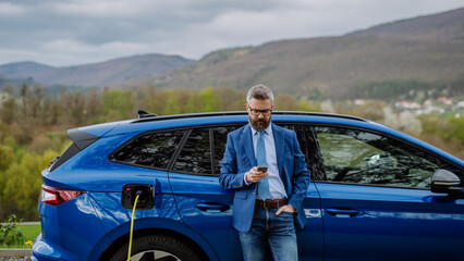 Wall Mural - Mature businessman waiting for charging his electric car.