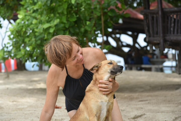 Happy attractive mature woman 50-60 and her dog sitting on the sand at the beach. Senior woman wear black swimsuit. Concept - vacation, retirement, active elderly people, friendship.