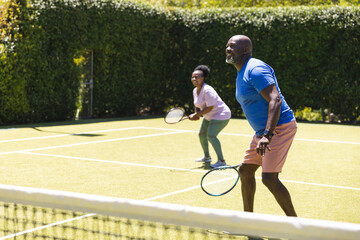 Happy senior african american couple playing tennis doubles on sunny grass tennis court, copy space