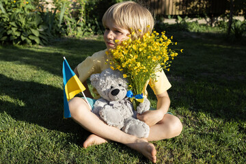 unhappy seven-year-old boy sits on grass with teddy bear, Ukrainian flag, bouquet of yellow flowers. Children against war.concept of peace for Ukrainian people. Loneliness, refugee, forced immigration