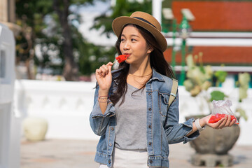 Portrait of Asian female traveler eating a slice of water melon while walking on sidewalk of buddhist temple on street in Bangkok, Thailand, Southeast Asia - Woman enjoying street food lifestyle