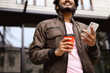 © Dmytro Hai - Smiling indian man in earphones holding coffee to go and using smartphone outdoors