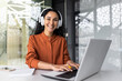 Happy and smiling hispanic businesswoman typing on laptop, office worker with curly hair and glasses, happy with achievement results, at work in office building, looking at camera in headphones.