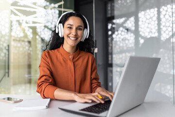 Wall Mural - Happy and smiling hispanic businesswoman typing on laptop, office worker with curly hair and glasses, happy with achievement results, at work in office building, looking at camera in headphones.