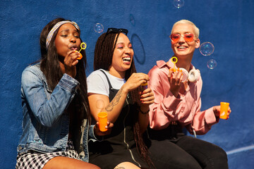 Happy, friends and women blowing bubbles on vacation, holiday travel or bonding activity and against a blue wall. Happy, game and ladies playing with soap or wand toy or in freedom and social break