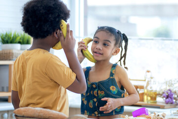 Cute little boy pretends to use a banana to talk on the smartphone, selective focus, boy and girl playing having fun in kitchen, imagination and creativity concept