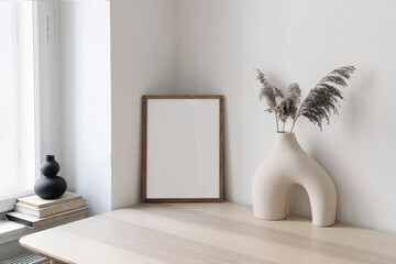 Vertical wooden picture frame, poster mockup in the corner. Wooden table, desk. Modern organic shaped vase. Dry flowers, grass. Old books on window sill. Home staging. Minimal Scandi boho interior.