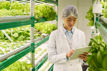 Aufkleber - Young serious Asian scientist in lab coat scrolling through online information in tablet while standing between vertical trusses