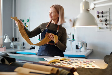 Aufkleber - Happy blond woman in workwear looking at piece of suede or leather in her hands while standing by workplace with working supplies