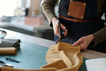 Aufkleber - Hands of mature craftswoman with scissors cutting piece of yellow suede or leather while working over new items for fashion collection