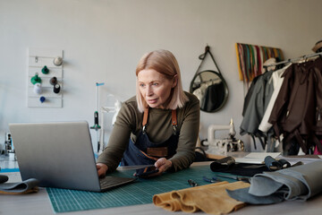 Aufkleber - Blond mature owner of leatherworking shop looking at laptop screen while standing by workplace and scrolling through new online orders