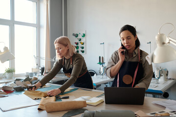Aufkleber - Young brunette female artisan using laptop and talking on smartphone by workplace while her mature colleague working with leather textile