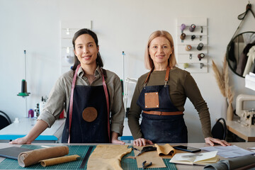 Aufkleber - Happy young and mature craftswomen in workwear looking at camera while standing by workplace with pieces of textile in workshop