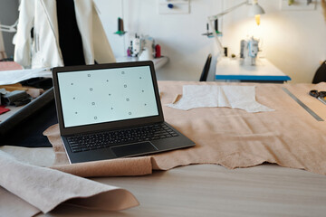 Poster - Workplace of tanner or taillor with laptop standing on beige suede or leather textile with paper patterns against sewing equipment