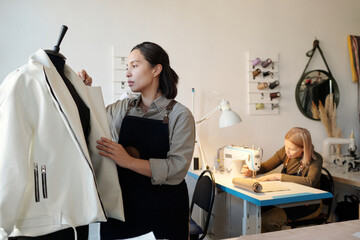Poster - Young brunette woman standing by mannequin with new white leather jacket against mature colleague sewing clothes on machine