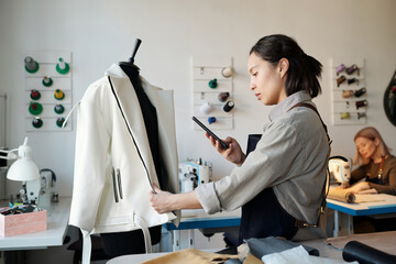 Aufkleber - Side view of young female fashion designer scrolling in smartphone or taking photo of new leather item hanging on mannequin