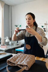 Aufkleber - Young craftswoman in apron taking photo of new handmade leather handbag on smartphone while standing by workplace in studio