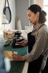 Poster - Young female leatherworker pressing small steel shaft down piece of leather or suede while going to hit it with hammer in workshop