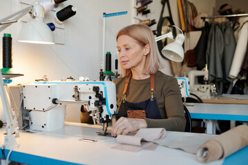 Aufkleber - Mature blond seamstress sitting by electric sewing machine during work over new seasonal fashion colleaction in workshop or studio