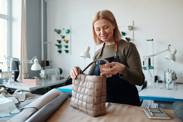Aufkleber - Happy mature female leatherworker looking at new beige handmade handbag while standing by workplace with textiles in craft shop
