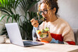 © simona - Woman busy working on laptop and eating a salads at the same time. Fast lunch break for online job activity. Concept of diet and modern work. Casual female in telecommuting smart working life alone