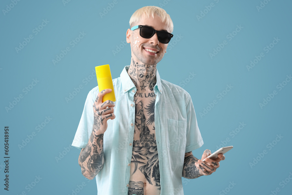 Tattooed man with phone and sunscreen cream on light blue background