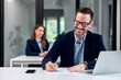 © bnenin - Smiling man dressed elegantly, working at the office, female colleague sitting in the background.