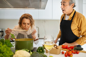 Elderly spouses cooking lunch together in kitchen, elderly wife trying meal while aged man cutting vegetable salad