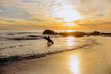 Sunset On The Beach On The Coast Of Southern California In The Winter With Silhouette Of Surfer Carrying Surfboard.