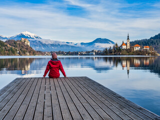 Wall Mural - A woman sitting on a wooden dock at lake Bled, enjoying the view of island church, Bled castle and the snow peak mountains, Slovenia