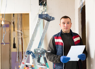 Wall Mural - Repair man standing in apartment at stepladder with documentation in hands and looking in camera.