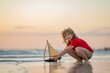 Little blonde boy put toy boat in the sea waves at the beach during summer vacation. Childhood and summer family vacation. Dream on travel. Travelling tour on sailing ship. Kids dream concept.
