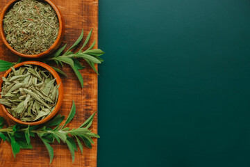 Wall Mural - Stevia dry leaves and powder in round wooden bowls and fresh plant branches on a wooden board on a dark green background