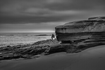 Wall Mural - 2021-07-01 A UNIDENTIFIED PERSON SITTING ON A BUCKET AT THE BEACH FISHING IN THE EARLY MORNIG BY A ROCKY OUT CROPPING IN LA JOLLA CALIFORNIA IN BLACK AND WHITE