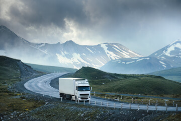 Large white transport truck transporting commercial cargo in semi trailer running on turning way highway road with scenic mountains mountaineous scenery in background.